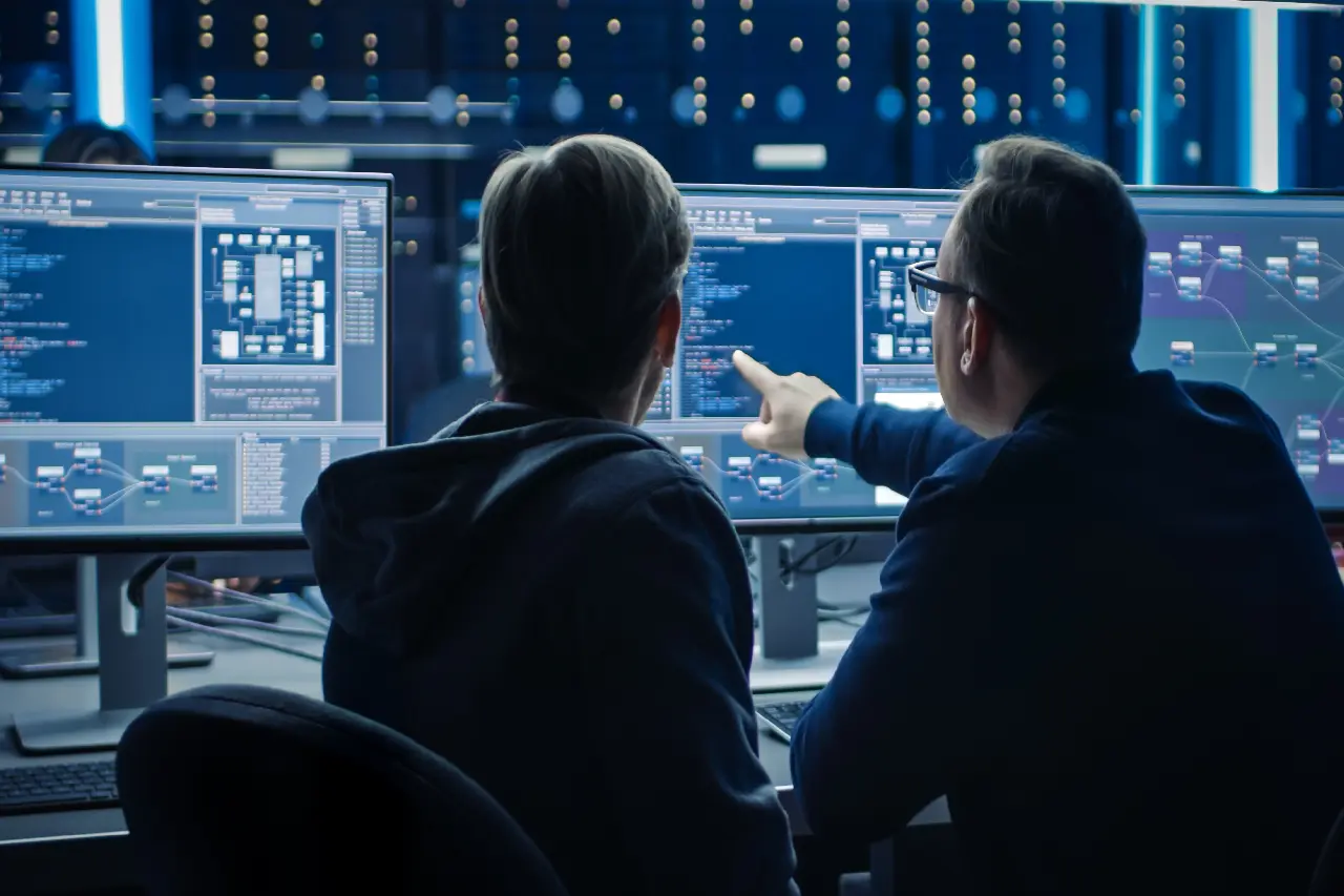 Two people sit at a desk, analyzing code on multiple computer screens in a dimly lit room. One person is pointing at the screen.