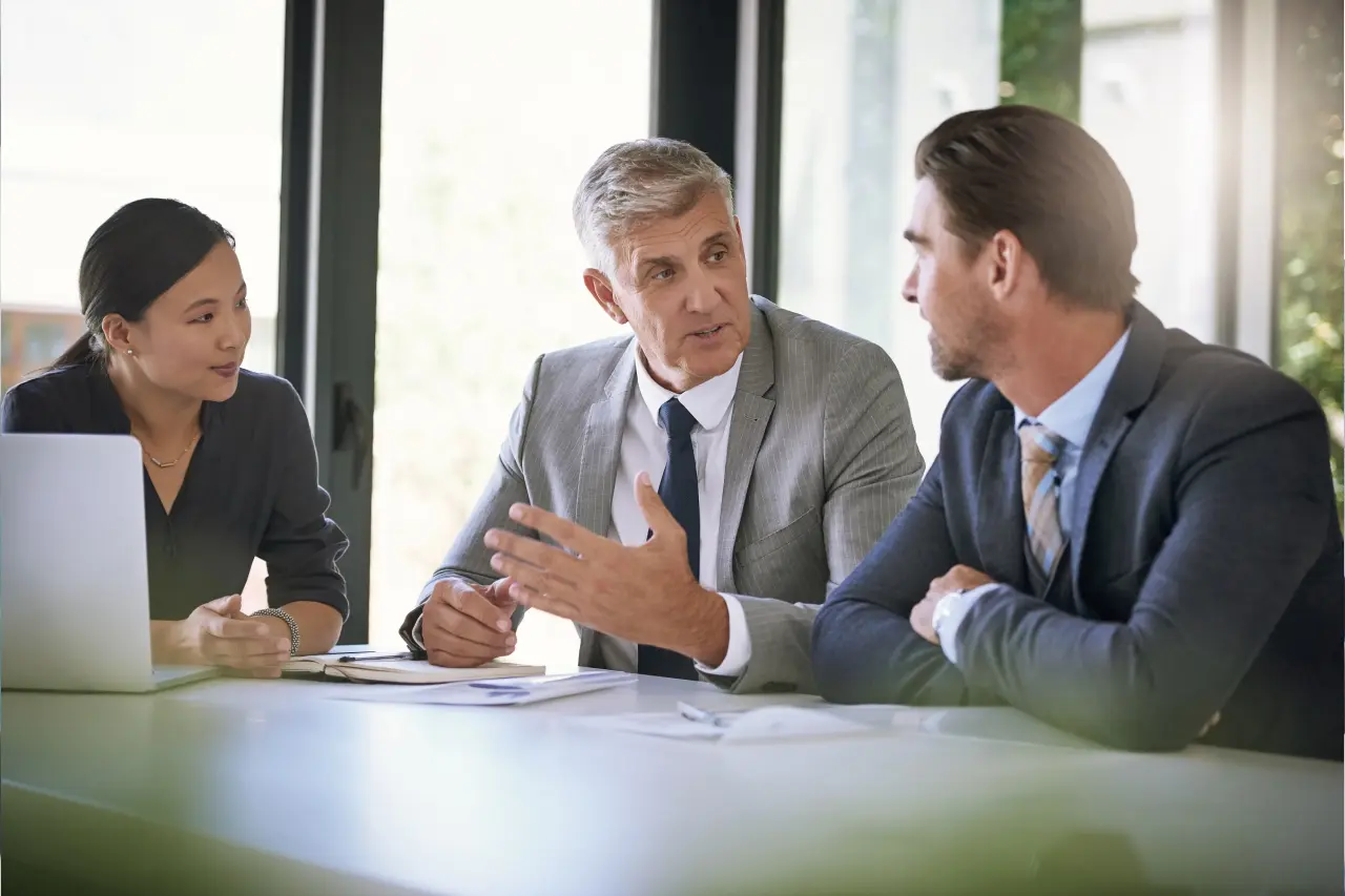 Three business professionals engage in discussion at a meeting table, with a laptop and documents.